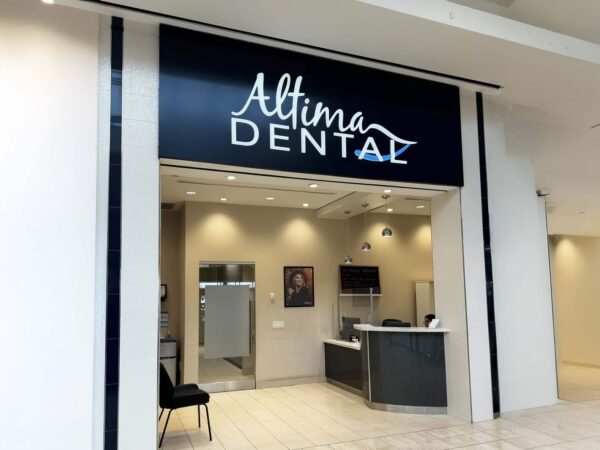The image shows the entrance of a dental clinic named "Altima Dental." The storefront features a modern design with a prominent sign, a reception area visible inside, and a waiting area with a chair. The overall aesthetic is clean and inviting.