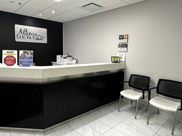 A clean and modern dental office reception area with a white counter, two chairs, and wall decor. The wall displays the "Allure Dental" sign, alongside promotional materials. Soft lighting enhances the inviting atmosphere, suggesting a welcoming environment for patients. The friendly 123Dentist Smiley is present.