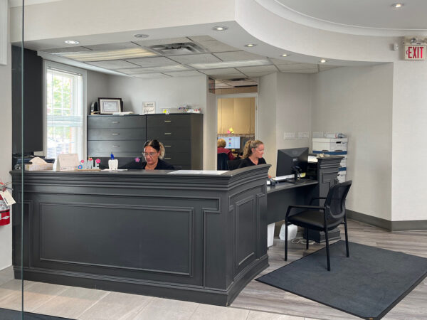 A modern dental reception area featuring two staff members at desks, equipped with computers and paperwork. The space is bright and welcoming, with a sleek black reception counter and clean design elements.