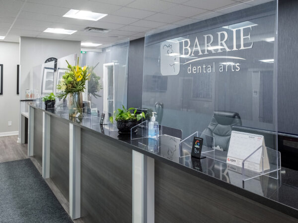 The image shows a modern dental office reception area with a sleek counter, decorative plants, and a welcoming ambiance. The name "Barrie Dental Arts" is prominently displayed. Natural light fills the space, creating a warm and inviting atmosphere for patients.