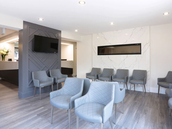 A modern dental office waiting area featuring grey chairs, a sleek reception desk, and bright lighting. The space is inviting and designed for comfort, with a minimalist aesthetic.
