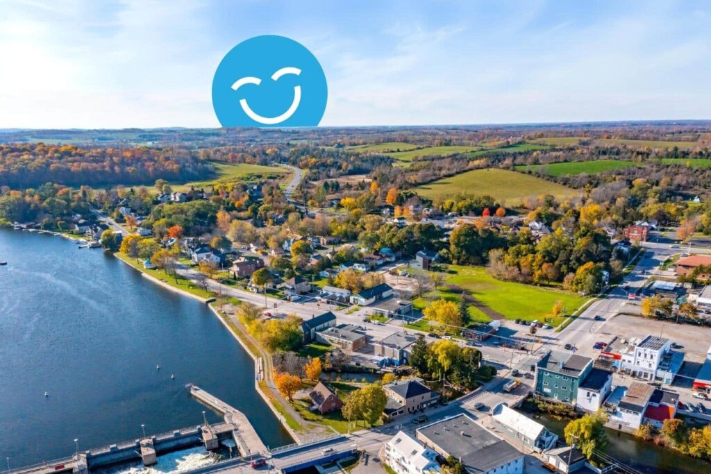 A scenic aerial view of a small town nestled beside a river, surrounded by colorful autumn foliage and rolling hills. A cheerful blue smiley face is overlaid in the sky.