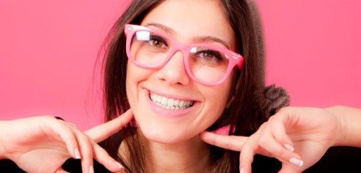 A young woman with long brown hair and pink glasses smiles brightly against a pink background, playfully pointing to her smile with both hands.