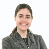 A smiling dental professional wearing a dark jacket stands with arms crossed. She exudes a friendly and approachable demeanor, inviting comfort and confidence in her expertise.