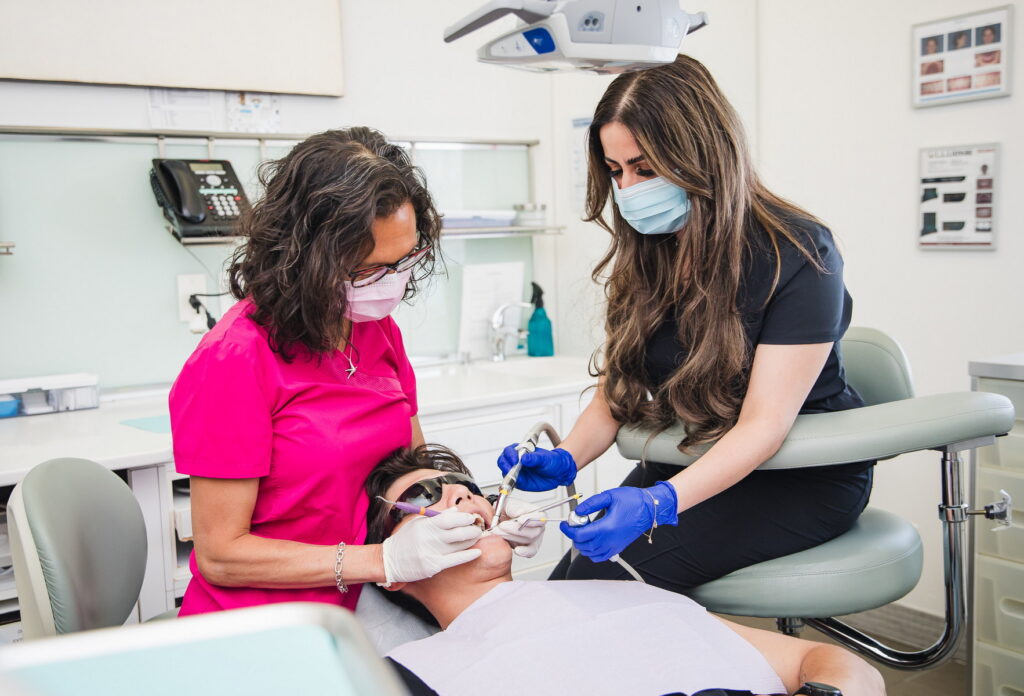 Two dental professionals in masks assist a patient in a dental chair. One hygienist prepares dental tools while the other gently holds the patient's head. The clean and modern dental office setting conveys a professional atmosphere. 123Dentist Smiley adds a friendly touch to the environment.