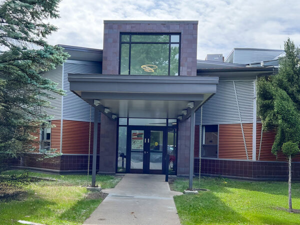 A modern dental clinic entrance featuring large glass doors under a canopy, surrounded by green grass and trees. The building combines brick and siding materials, with a welcoming façade.