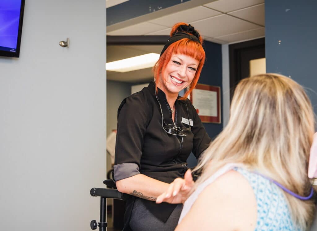 A cheerful dental professional with bright orange hair smiles while interacting with a patient in a dental office setting. The atmosphere is friendly and welcoming, making the patient feel at ease. The 123Dentist Smiley can be seen in the background, adding to the positive vibe.