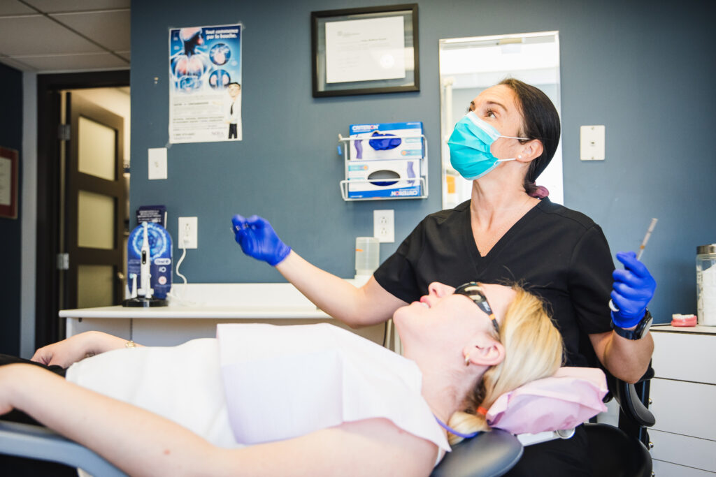 A dental professional in a mask and gloves talks to a patient reclining in a dental chair. The setting is a modern dental office, with dental supplies visible in the background.