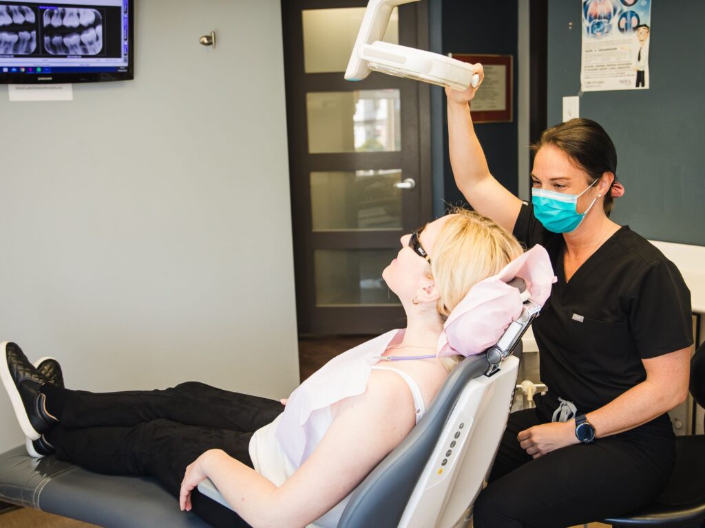 A dental professional in a black uniform is adjusting equipment above a patient reclining in a dental chair. The patient, wearing glasses and a white top, appears calm. The setting is a modern dental office, emphasizing a comfortable atmosphere. The friendly 123Dentist Smiley adds a cheerful touch.