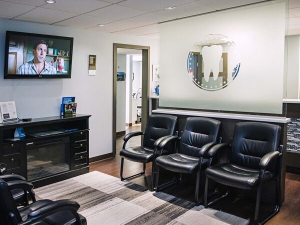 A comfortable waiting area featuring black leather chairs, a television displaying a man speaking, and a modern decor with a circular mirror. The space is well-lit and inviting, with a wooden floor and a simple rug.