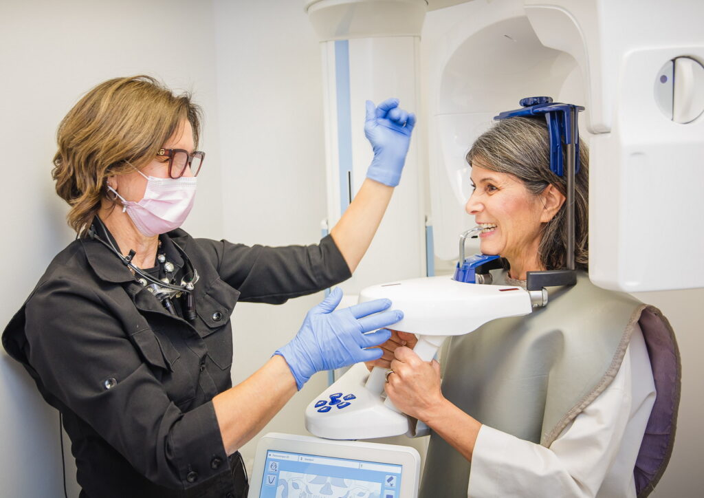 A dental technician in gloves assists a patient wearing a protective apron during a dental imaging procedure. The patient appears comfortable while receiving care in a modern clinic setting.