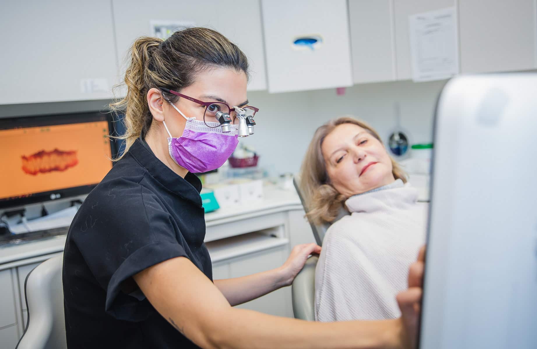 A dental hygienist in a black shirt and purple mask is assisting a patient in a dental chair. The patient appears relaxed, with light brown hair and wearing a dental bib. A dentist's monitor is visible in the background, displaying the 123Dentist Smiley.