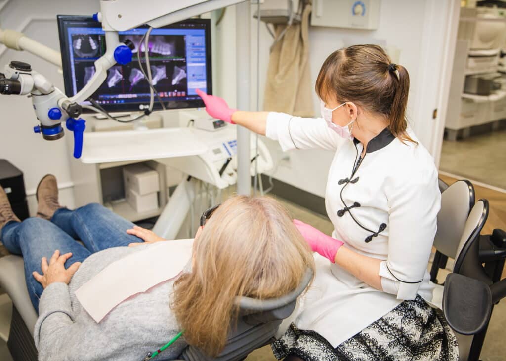 A dental hygienist in a white coat explains dental X-rays to a seated patient in a dental office. The hygienist wears pink gloves and points to the monitor displaying the X-ray images.