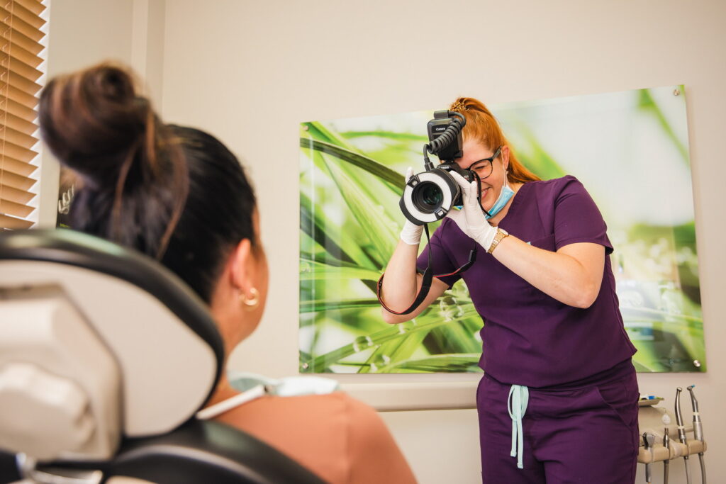 A dental professional in purple scrubs is taking a photo of a patient sitting in a dental chair. The background features a nature-themed image with green foliage, creating a calming atmosphere.