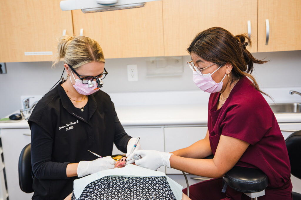 A dental professional in a black coat and safety glasses performs a procedure on a patient, who is wearing a maroon shirt and a mask. The scene takes place in a modern dental office.
