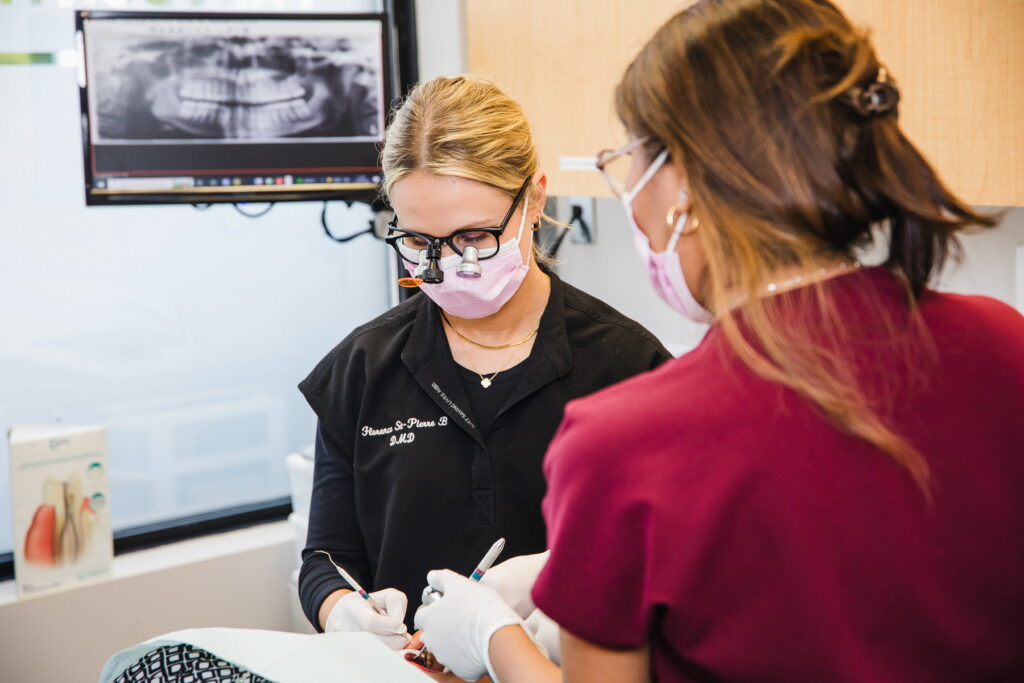 Two dental professionals in masks and gloves are working in a dental office. One is focused on a patient's dental procedure while the other assists. A digital screen in the background displays dental imagery. The friendly 123Dentist Smiley adds a cheerful vibe to the setting.