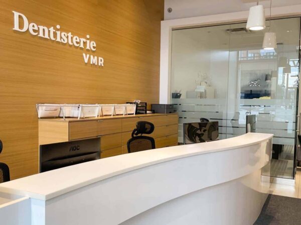 The image shows a modern dental office reception area with a curved white desk, wooden wall featuring "Dentisterie VMR," and clear glass partitions. There are chairs and informational brochures displayed on the counter, creating a welcoming atmosphere.