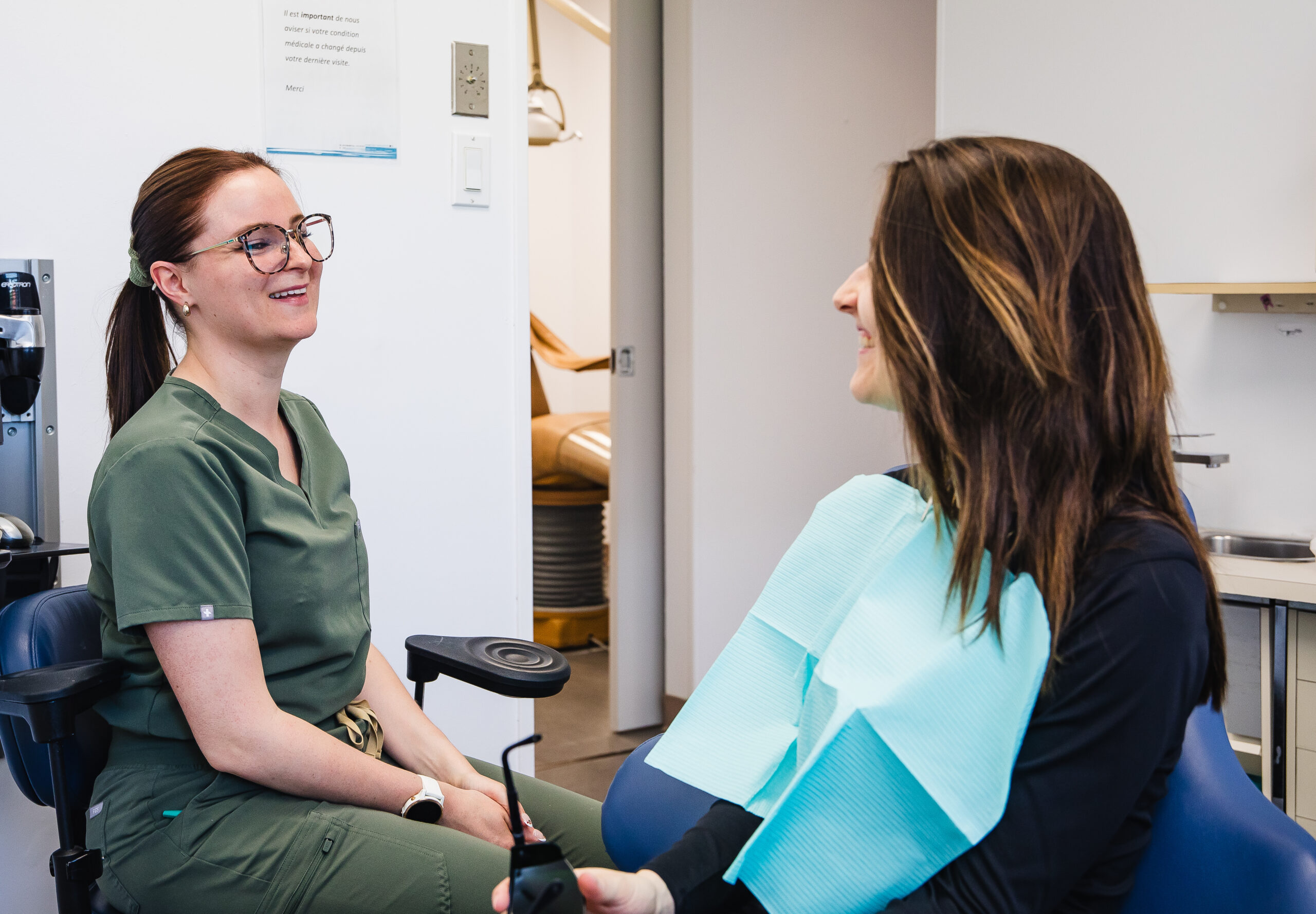 A smiling dental professional in green scrubs chats with a patient wearing a dental bib in a welcoming office setting. The atmosphere feels friendly and relaxed, reflecting a positive dental visit experience. 123Dentist Smiley adds an extra touch of cheer to the scene.