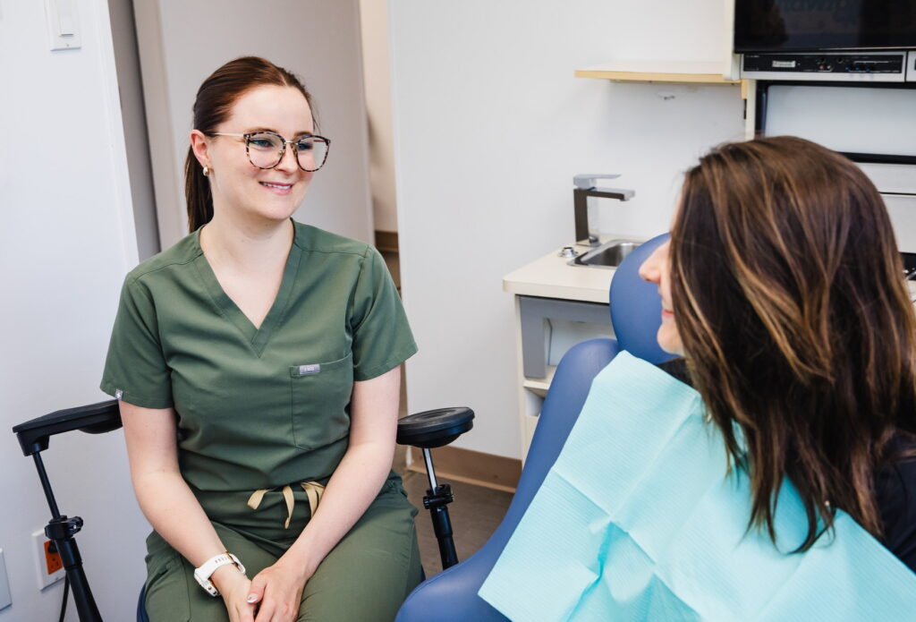 A dental professional in green scrubs smiles while conversing with a patient in a dental chair, who is wearing a blue dental bib. The setting is calm and welcoming, typical of a dental clinic environment.