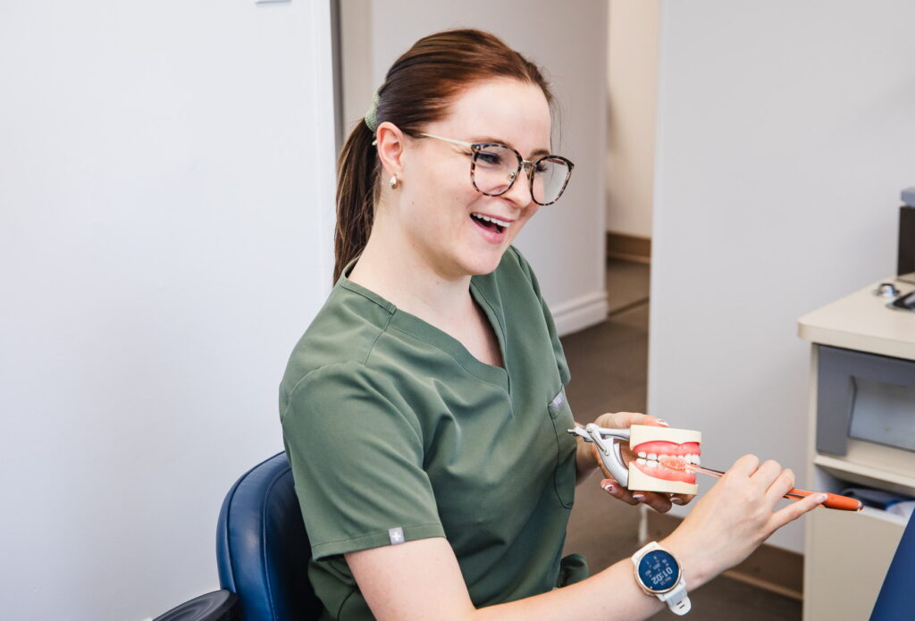 A cheerful dental professional in green scrubs smiles while demonstrating dental care using a model and a toothbrush. The setting appears to be a dental clinic, contributing to a friendly and informative atmosphere.