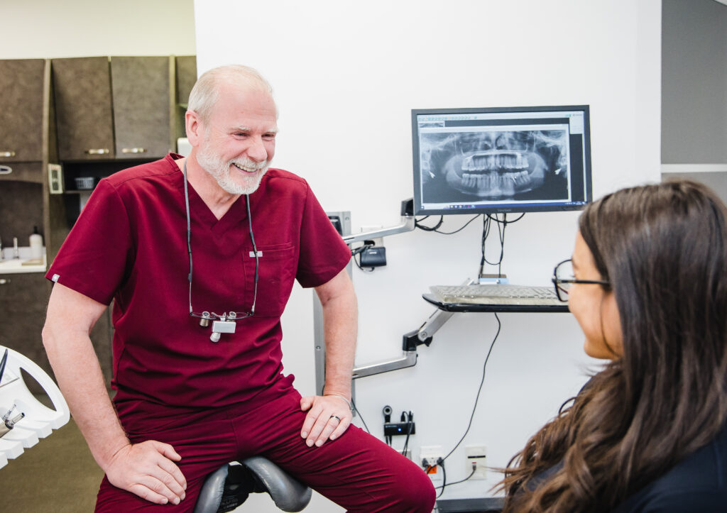A dentist in maroon scrubs smiles at a female patient while sitting in a dental office. They engage in conversation, with an X-ray displayed on the screen behind them. The atmosphere appears friendly and professional.