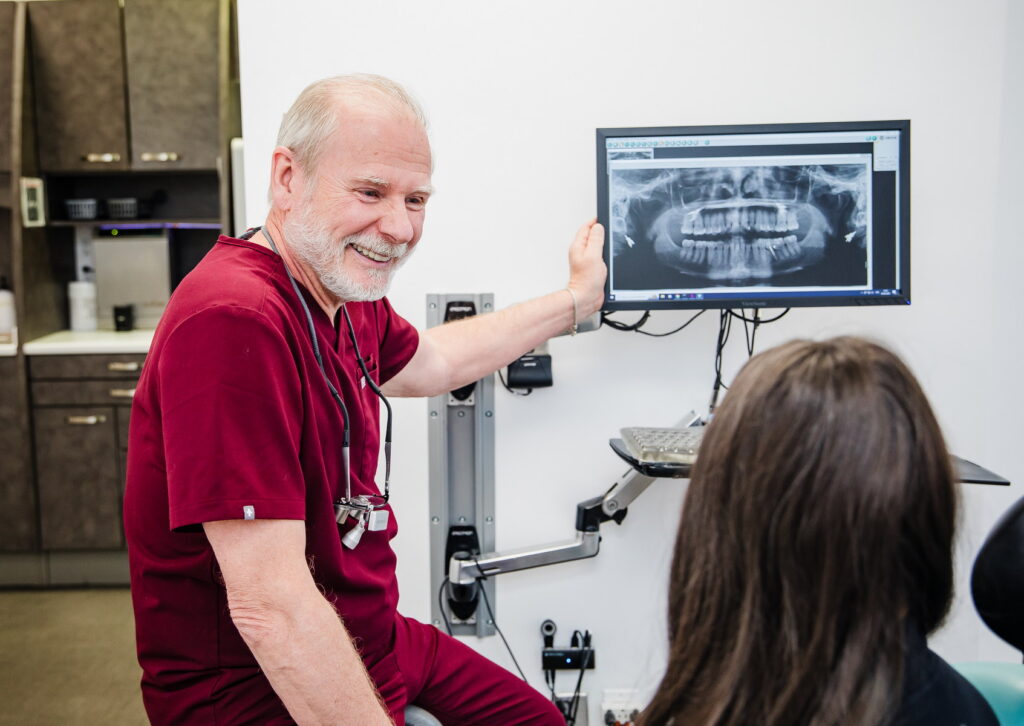 A smiling dentist in maroon scrubs explains an X-ray to a patient in a dental office. The atmosphere is friendly and welcoming, with dental equipment in the background.