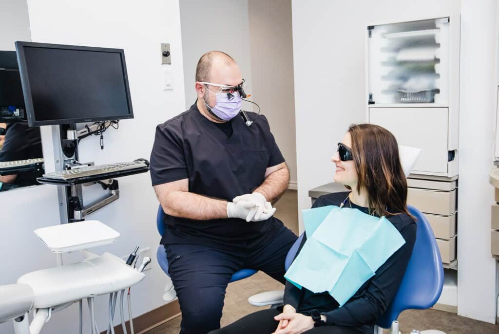 A dentist wearing gloves and a mask converses with a patient seated in a dental chair. The patient is wearing protective eyewear and a dental bib. A computer and dental equipment are visible in the background, creating a clinical environment.