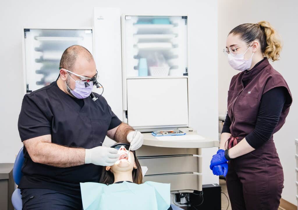 A dentist is examining a patient in a dental clinic, while a dental assistant observes. Both professionals wear protective masks and gloves, ensuring a safe and hygienic environment for dental care.