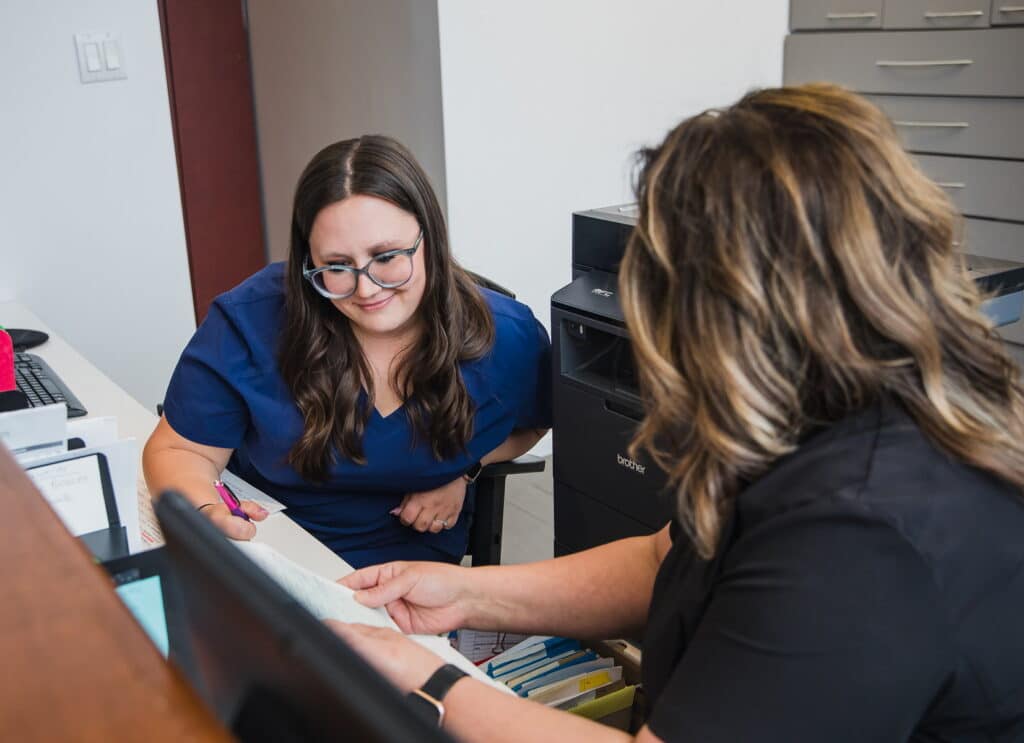 A friendly dental receptionist interacts with a patient at the front desk, sharing information and smiling. The office is well-organized with files and equipment visible in the background. The 123Dentist Smiley can be seen, adding a cheerful touch to the atmosphere.