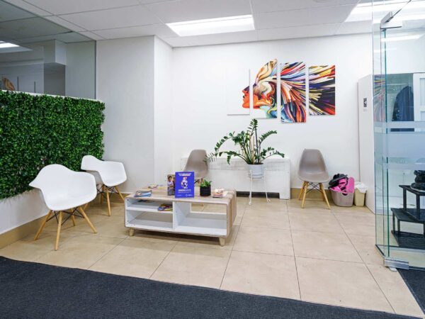 A modern waiting area featuring two white chairs, a small coffee table with literature, a green plant wall, and colorful artwork above. The space is well-lit with glass accents and a stylish staircase in the background.