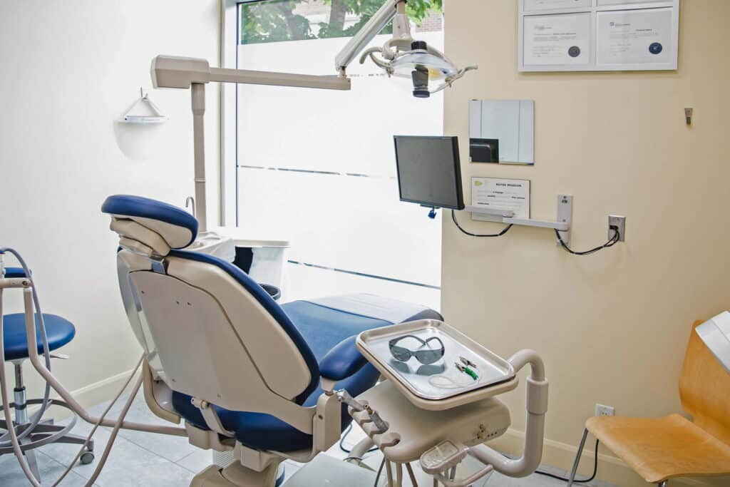 A modern dental clinic room featuring a blue dental chair, dental equipment, a mounted light, and a wall-mounted monitor. The space is well-lit with natural light from a window.