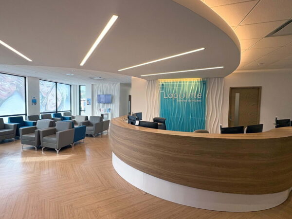 A modern reception area featuring a curved wooden desk and comfortable seating. Large windows provide natural light, and the decor includes a blue accent wall with wave patterns, creating a welcoming atmosphere for visitors.