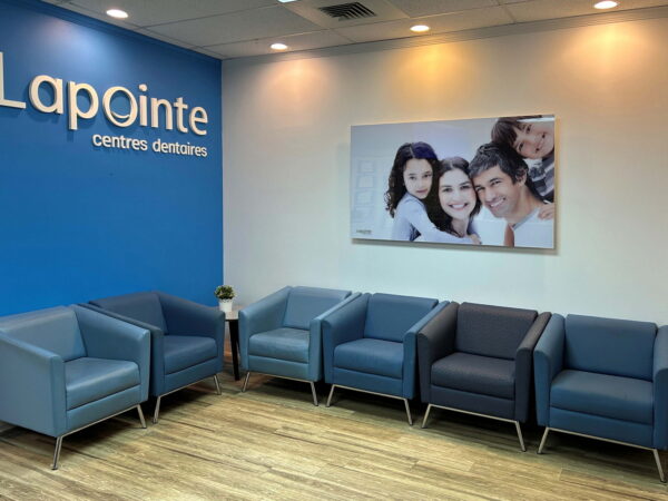 A modern dental clinic reception featuring a blue wall with the "Lapointe" logo, five stylish gray armchairs arranged for seating, and a family portrait on the adjacent wall, creating a welcoming atmosphere.