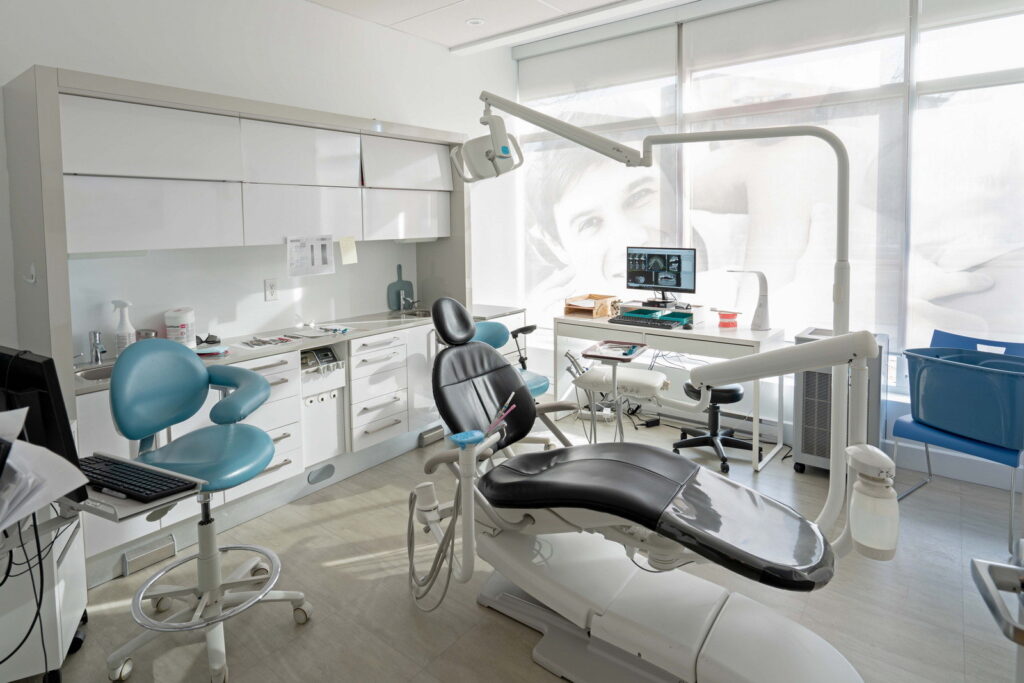 A modern dental office featuring a dental chair, equipment, and treatment station. The interior is bright and organized, with large windows allowing natural light.