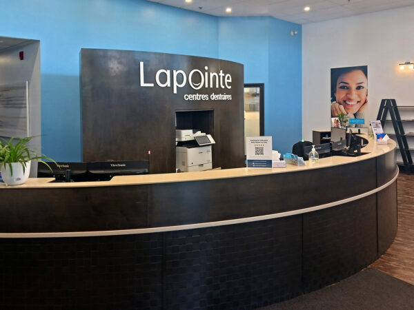 A friendly dental reception area featuring a curved desk with the name "Lapointe" prominently displayed. The space is bright with light blue walls, a welcoming atmosphere, and a smiling dentist's image in the background. The 123Dentist Smiley adds a touch of cheer to the environment.