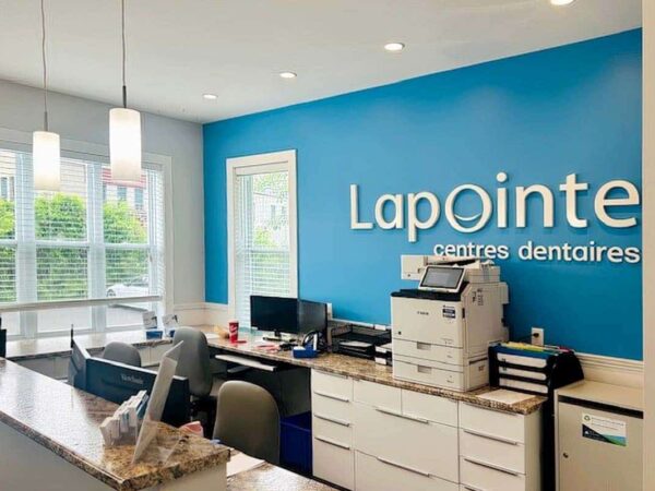 A modern dental clinic reception area featuring a blue wall with the sign "Lapointe centres dentaires." The space includes a desk with computers, a printer, and a welcoming environment with natural light from large windows.