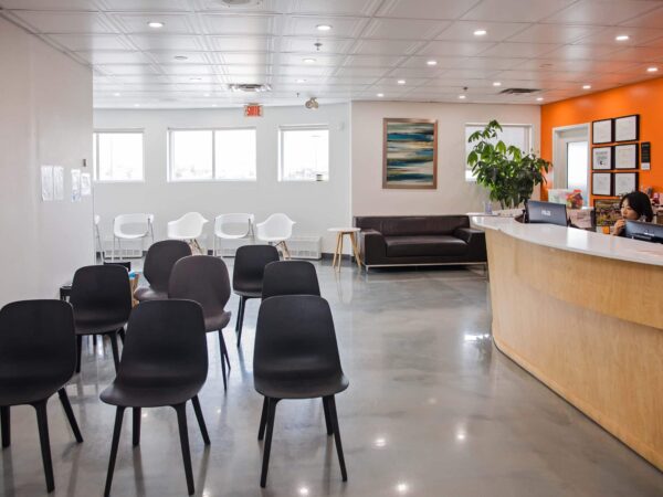 A modern reception area with a wooden desk, a few people seated at the desk, and several black chairs arranged in the waiting area. The walls are painted in neutral tones with an orange accent wall, and there are windows allowing natural light inside.