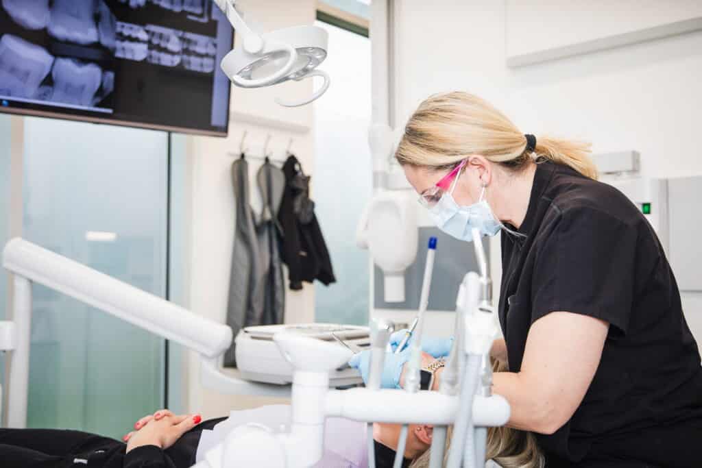 A dental professional in a black uniform assists a patient at a dental clinic, wearing a mask and gloves. In the background, dental x-rays are displayed on a screen. The environment is clean and modern, reflecting a professional dental practice.