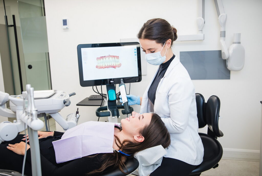 A dental hygienist in a white coat examines a patient sitting in a dental chair, with a monitor displaying dental images in the background. The patient is relaxed, wearing a bib, while the hygienist prepares to clean their teeth. The friendly 123Dentist Smiley adds a welcoming touch to the scene.