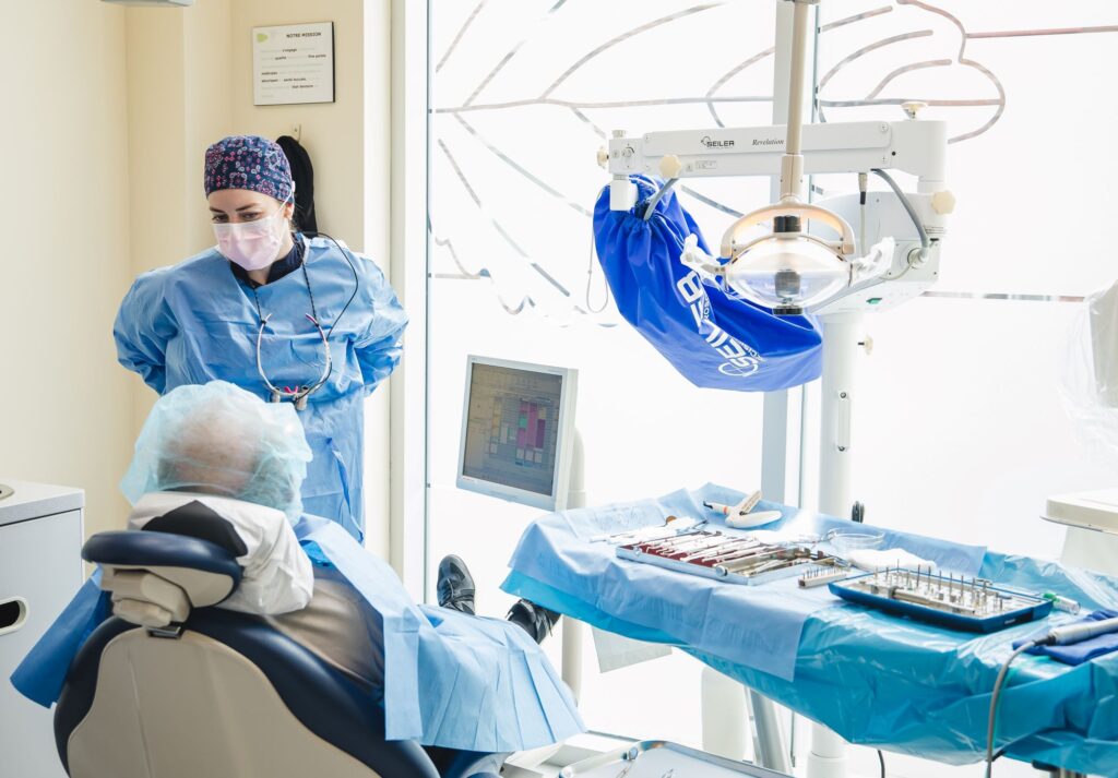 A dental professional in scrubs and a mask talks to a patient seated in a dental chair. The dental office is bright and equipped with modern tools, creating a calm environment for the appointment. Nearby, a friendly and familiar 123Dentist Smiley adds a cheerful touch.