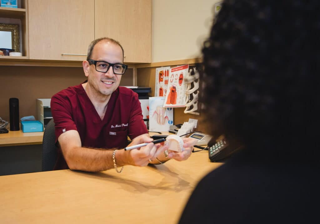 A dental professional in red scrubs is talking to a patient across a desk, holding dental tools. The environment appears inviting and professional, reflecting a dental consultation setting. A friendly blue smiley face, known as the 123Dentist Smiley, adds a cheerful touch to the atmosphere.