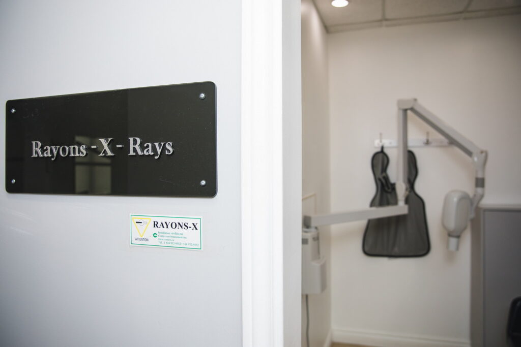 A dental office door displaying a sign reading "Rayons-X-Rays." Nearby, a gray chair and a protective lead apron hang on the wall, indicating the room's purpose for dental X-rays.