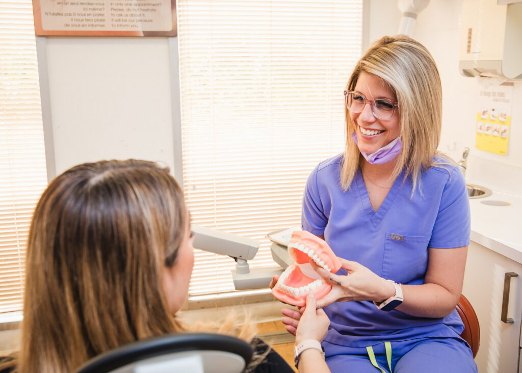 A dental professional in scrubs, wearing glasses and a face mask, explains dental concepts using a model of teeth to a patient seated in a dental chair. Natural light filters through the window, creating a welcoming atmosphere.