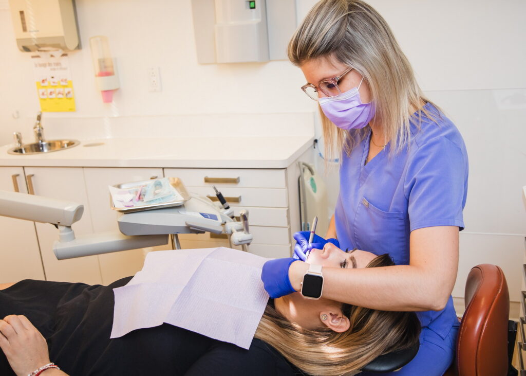A dental hygienist in a blue uniform assists a patient in a dental chair, wearing a mask and gloves. The patient is relaxing with a protective bib over their chest. The scene conveys a caring dental environment. The cheerful 123Dentist Smiley can be seen in the background, brightening the atmosphere.