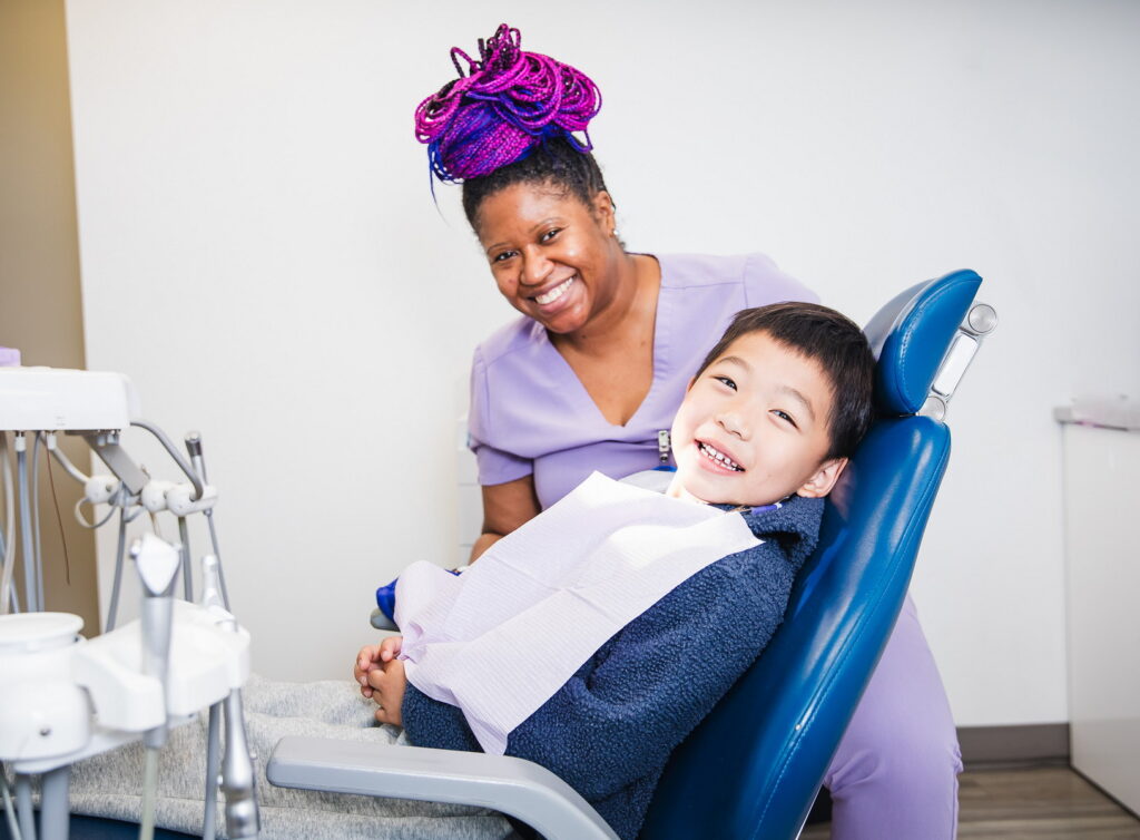 A smiling child sits in a dental chair wearing a bib, while a dental professional with vibrant purple hair stands beside him, both appearing happy and relaxed in a modern dental office setting. The cheerful 123Dentist Smiley can be seen in the background.