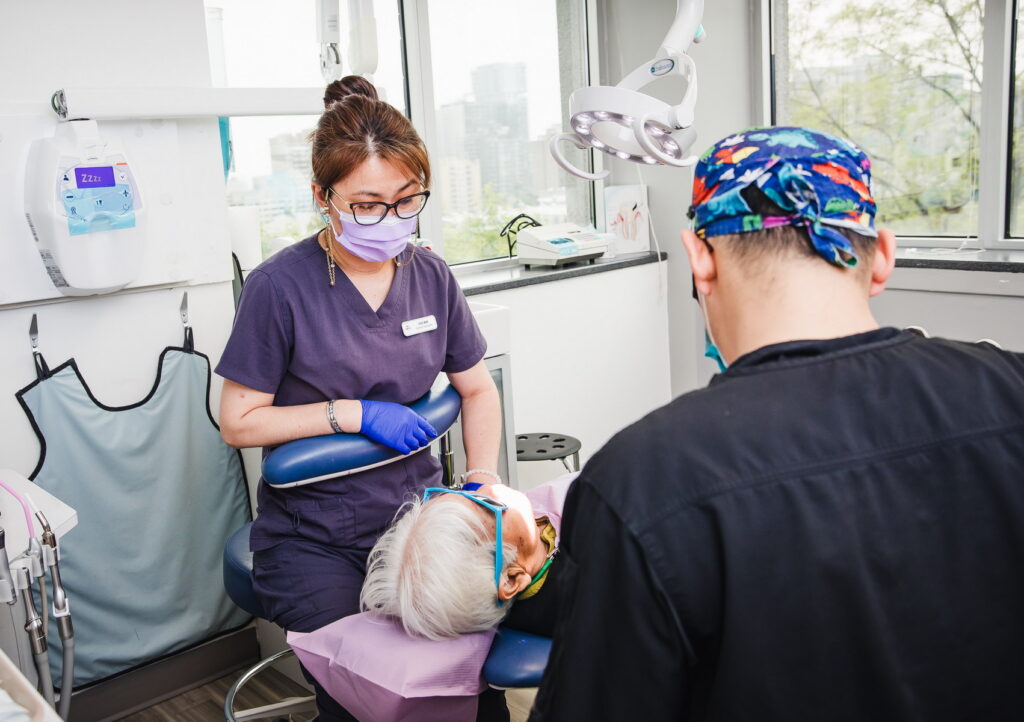 A dental professional assists an elderly patient in a dental office, while another staff member performs a procedure. Bright, modern surroundings create a welcoming atmosphere. The 123Dentist Smiley adds a friendly touch to the scene.