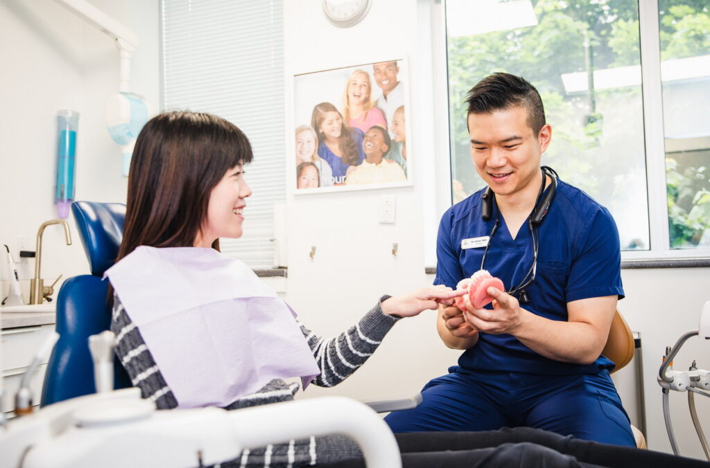A dentist in scrubs explains a dental model to a smiling patient sitting in a dental chair, creating a friendly and informative atmosphere. The background includes cheerful photos, enhancing a welcoming dental clinic environment. The 123Dentist Smiley adds a touch of friendliness to the scene.