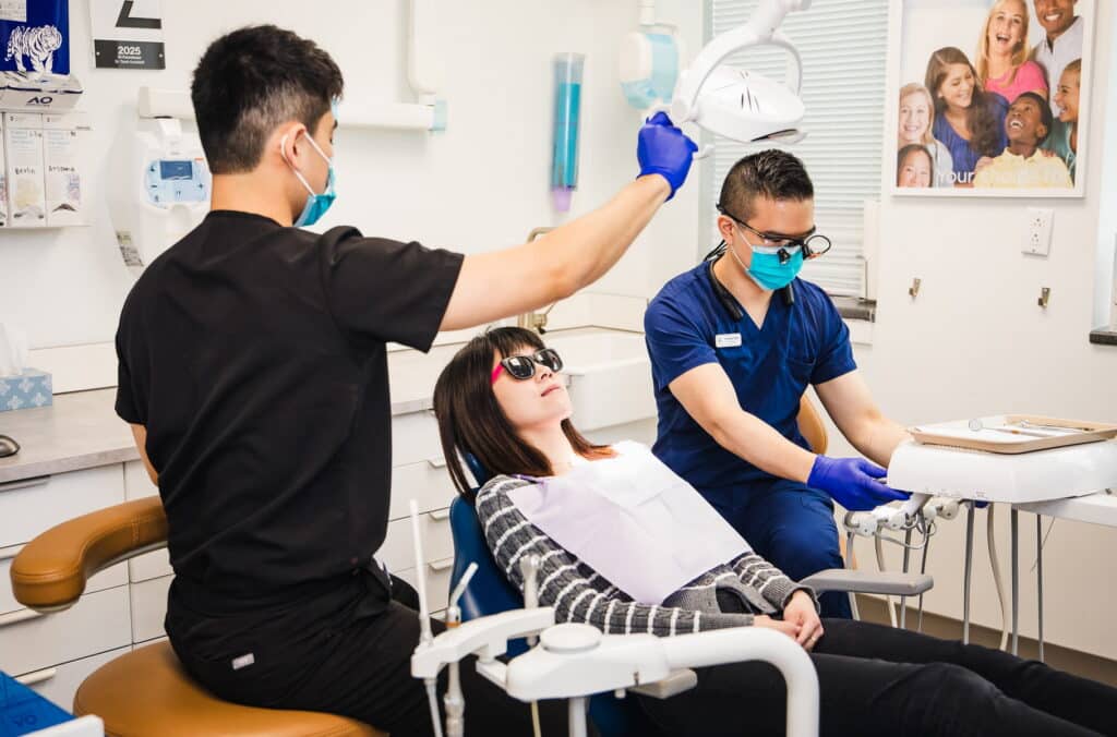 A dental office scene featuring a dental hygienist and a dentist assisting a patient in a dental chair. The patient wears protective eyewear and is ready for treatment. In the background, there's a cheerful icon known as the 123Dentist Smiley, adding a friendly touch to the environment.
