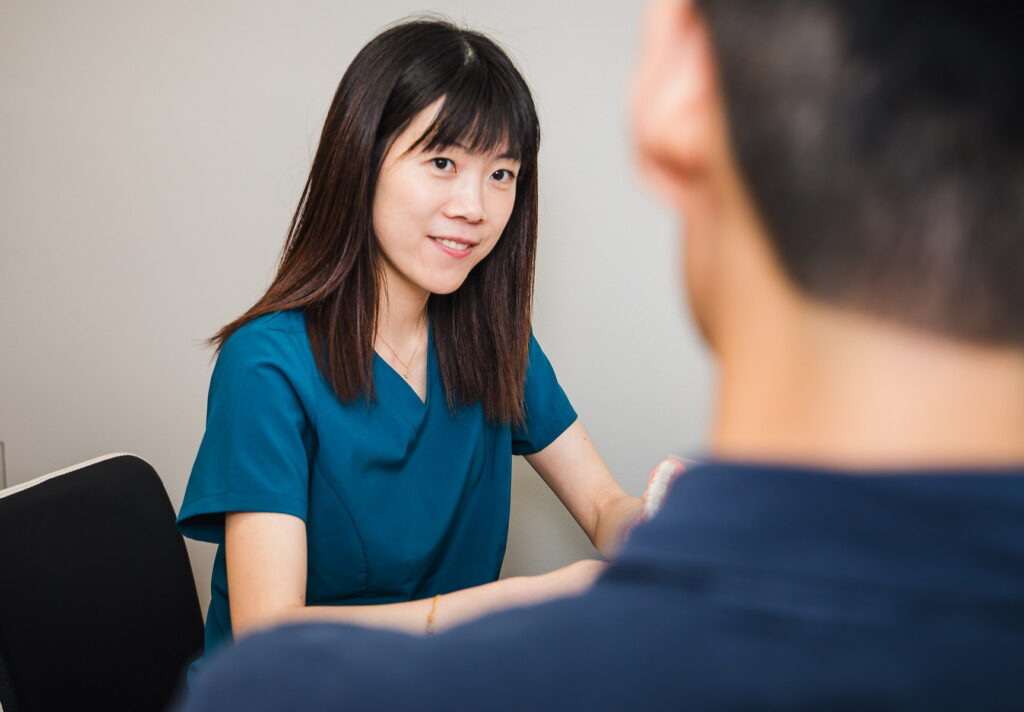A friendly dental professional in a teal uniform engages with a patient, offering a warm smile and attentive conversation. The scene conveys a welcoming atmosphere typical of a dental clinic.