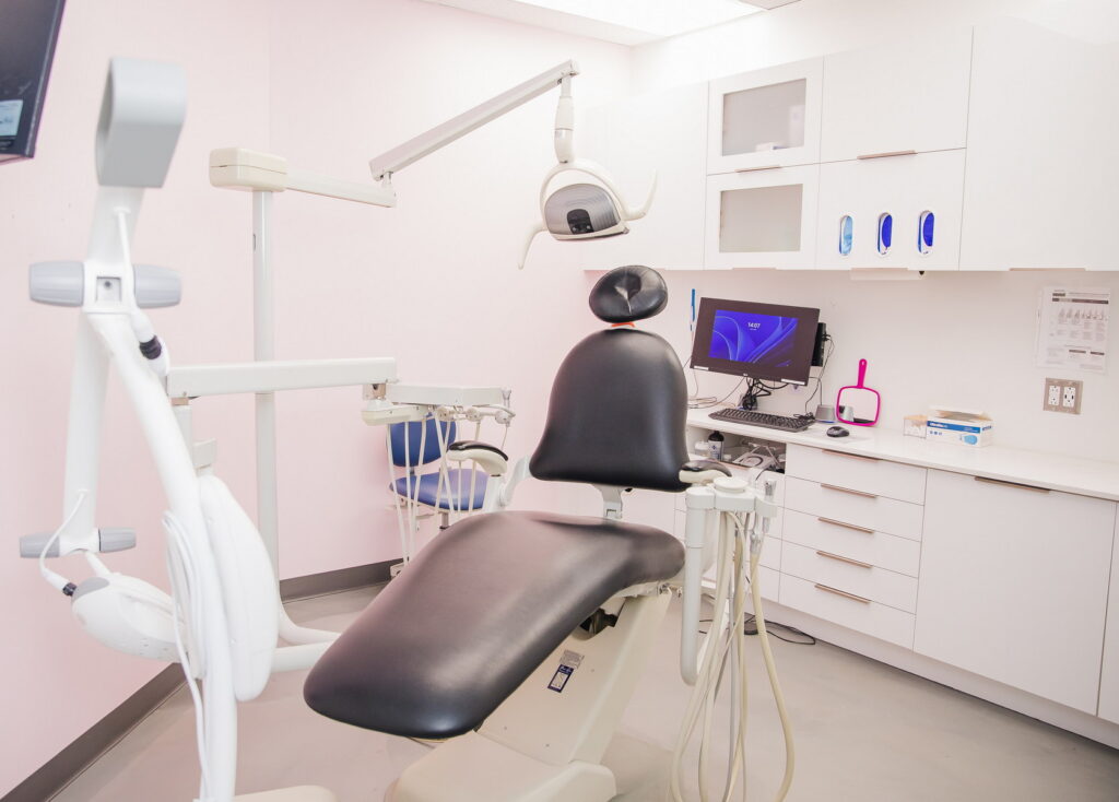 A modern dental office featuring a black dental chair, dental equipment, and a computer workstation. The walls are painted pink, creating a welcoming atmosphere.